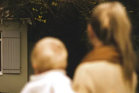 A woman and a child stand together in front of a house, showcasing a moment of connection and family warmth.