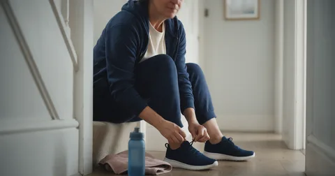 Person sitting on a bottom stair in a bright hallway, lacing a navy trainer, with a water bottle nearby.