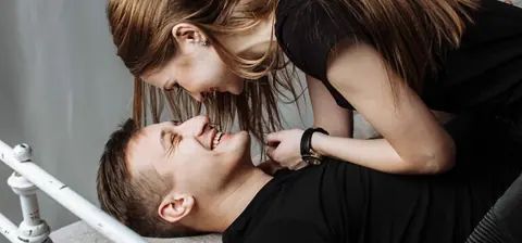 A smiling woman leans over a laughing man lying on a bed, their faces close together.