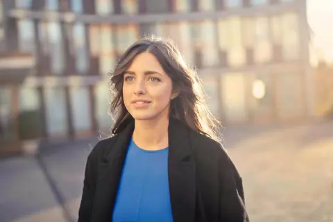 Smiling woman with shoulder-length brown hair and a black coat, standing outside in low sunlight
