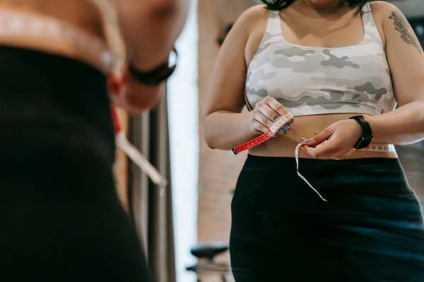 what is obesity | woman looking in mirror with measuring tape around waist